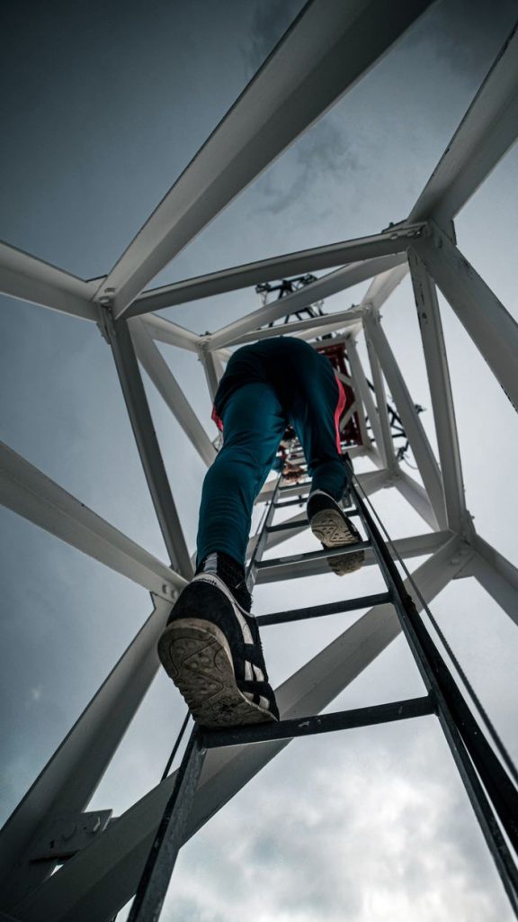 Person climbing a communication tower ladder against sky, emphasizing height and industrial aspect.